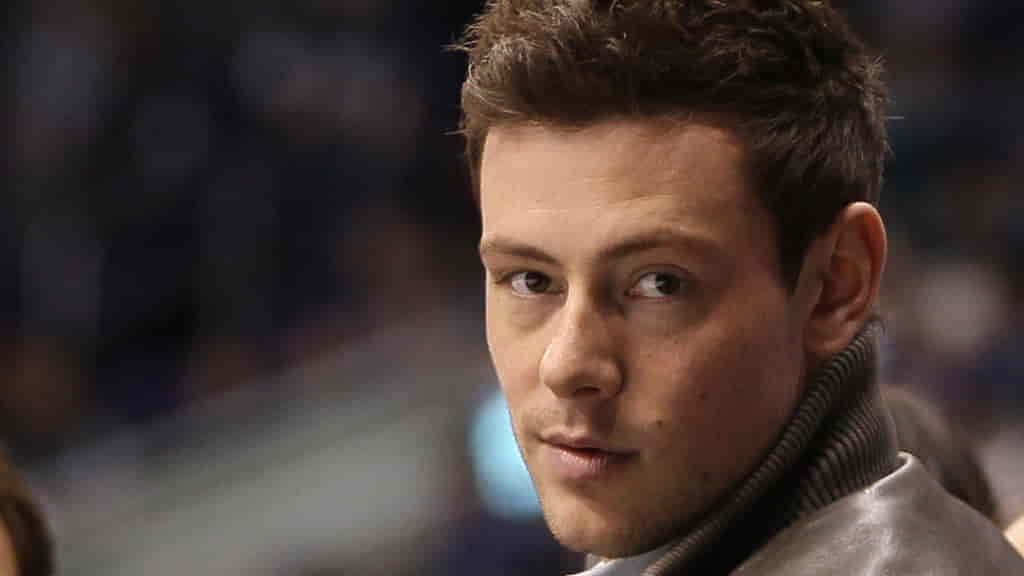 Close-up of Cory Monteith in a gray leather jacket and turtleneck sweater, gazing thoughtfully over his shoulder with a soft background blur of a public venue.