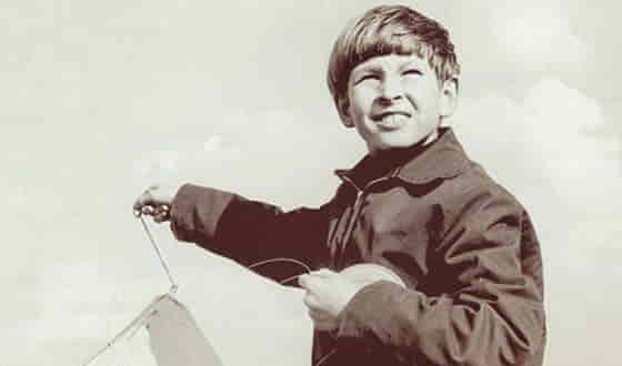 A young Lars von Trier smiling while flying a kite, dressed in a dark coat, under a cloudy sky in a vintage-styled photograph.