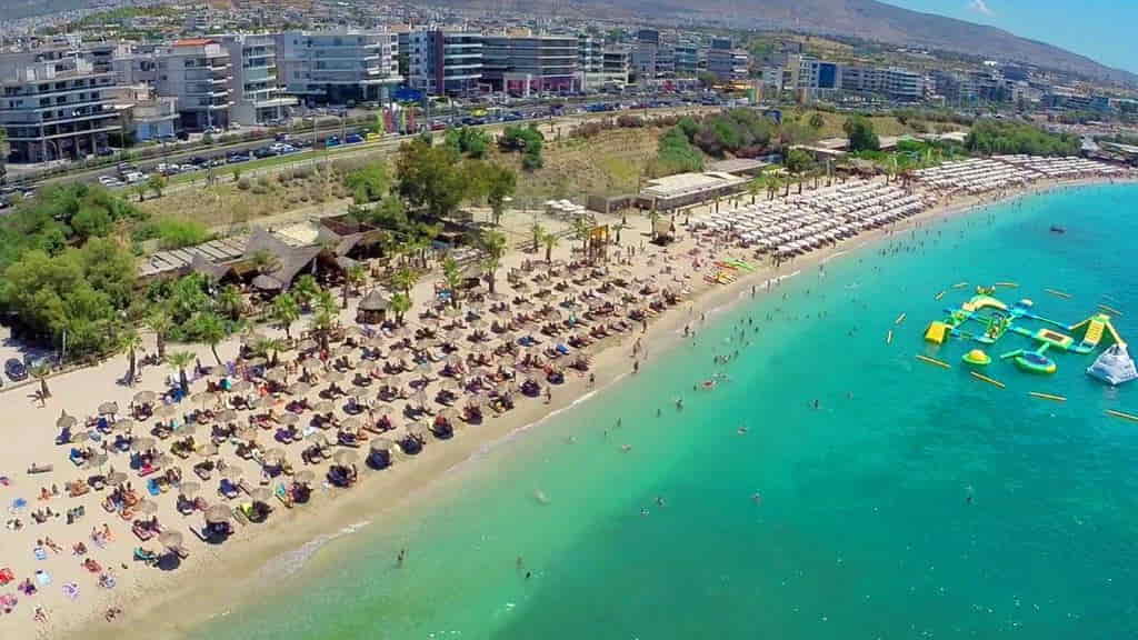 Aerial view of Edem Beach in Athens, Greece, showing turquoise waters, rows of umbrellas and sunbeds, beachgoers along the shore, and a floating water park near the coastline.