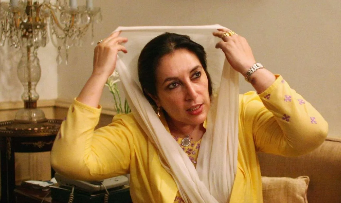 Benazir Bhutto speaking passionately at a public event, wearing a traditional outfit and gesturing with her hand.