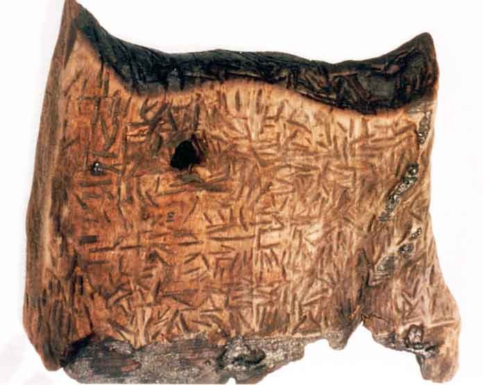 The Dispilio Tablet exhibited in a museum case, featuring visible carved symbols on its wooden surface from a Neolithic lakeside settlement in Greece.